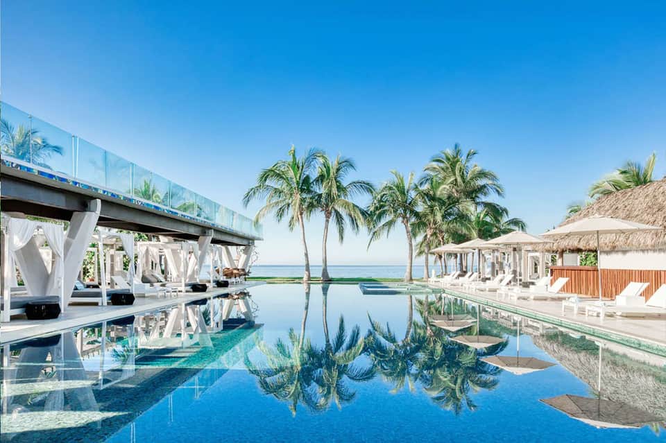 Infinity pool reflecting palm trees and ocean with white cabanas and covered lounging areas