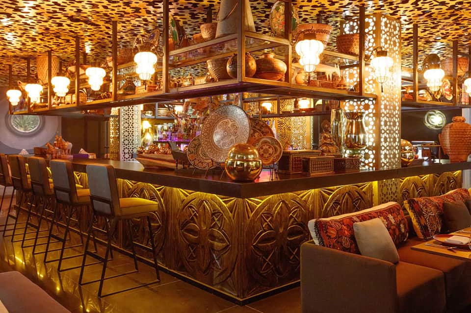 Upscale bar interior with ornate carved wood counter, hanging lights, decorative plates, and warm golden lighting