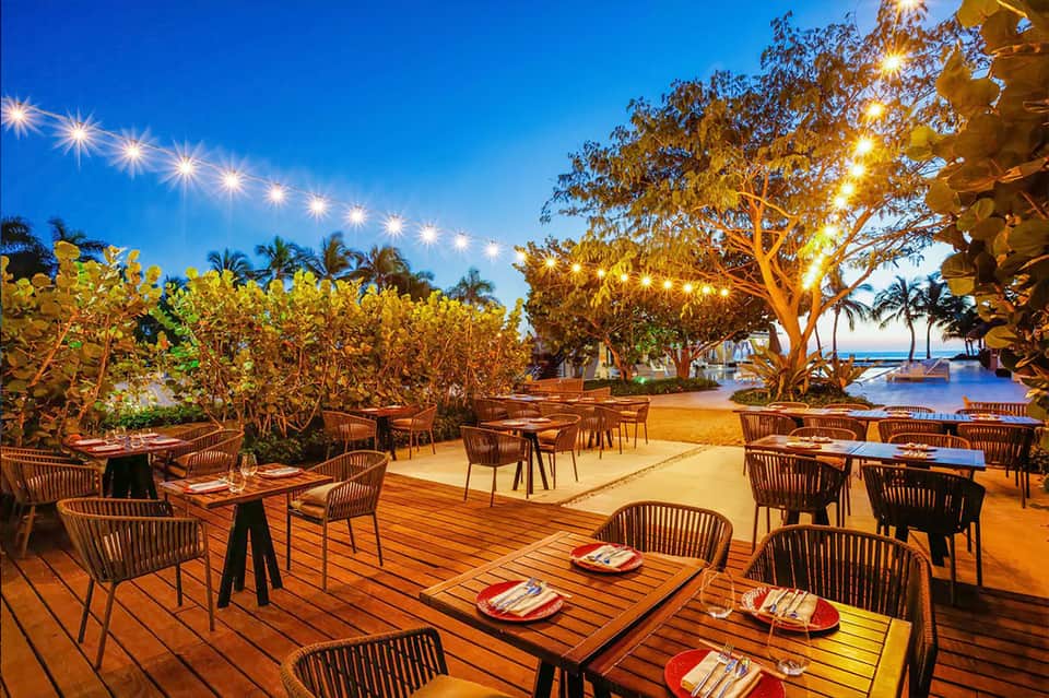 Beachfront restaurant terrace at dusk with woven chairs, wooden tables, and twinkling string lights