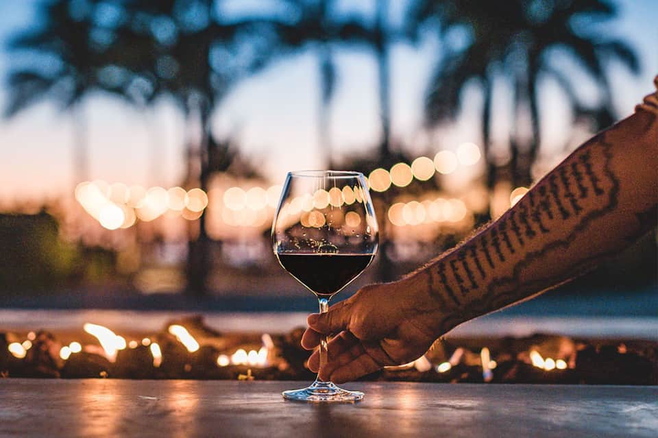 Hand holding wine glass at sunset with palm trees and string lights in background