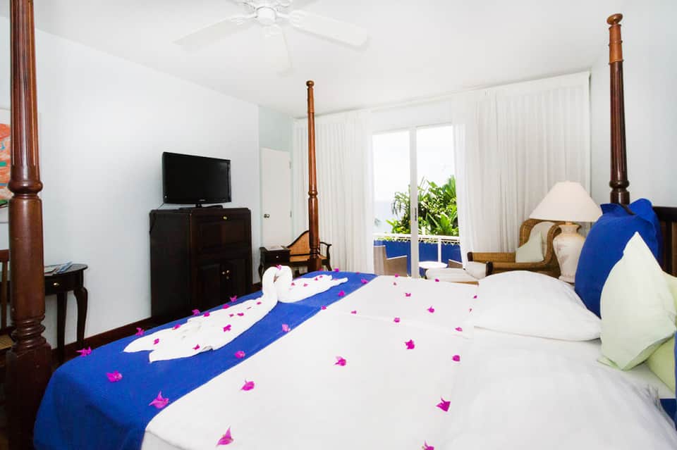 Resort bedroom with four-poster wood bed, blue decorative runner with pink flowers, ocean view from balcony.