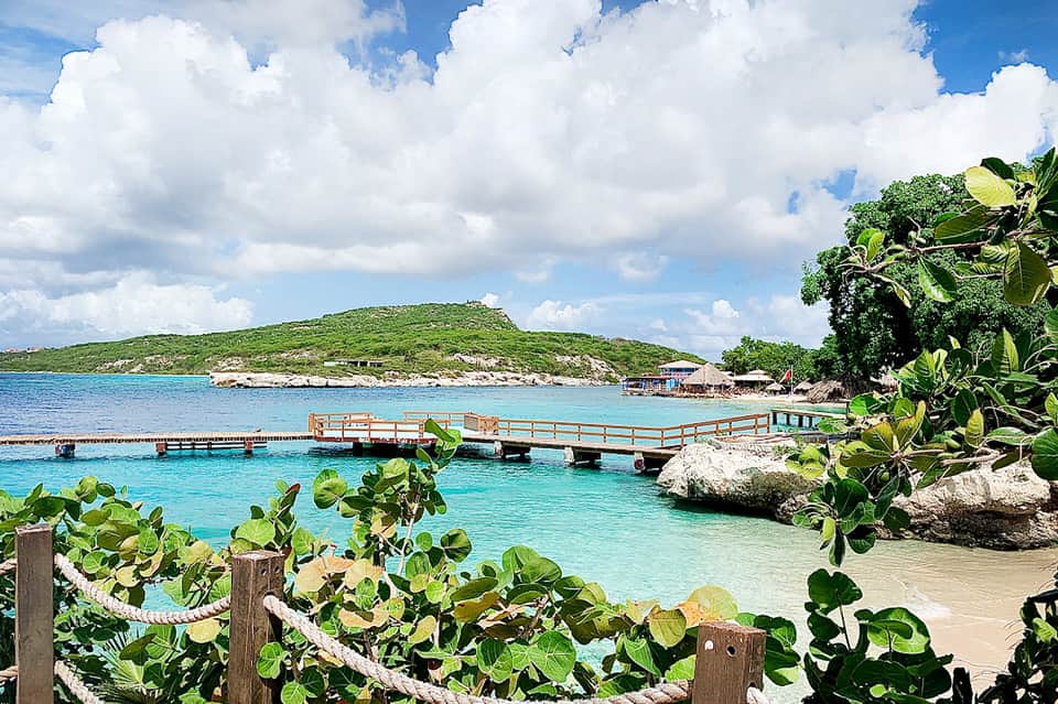 Tropical beach cove with turquoise water, wooden dock, green vegetation, and verdant hillside backdrop