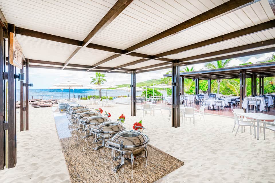 Beachfront dining area with chafing dishes under wooden pergola, ocean view, and lounge seating