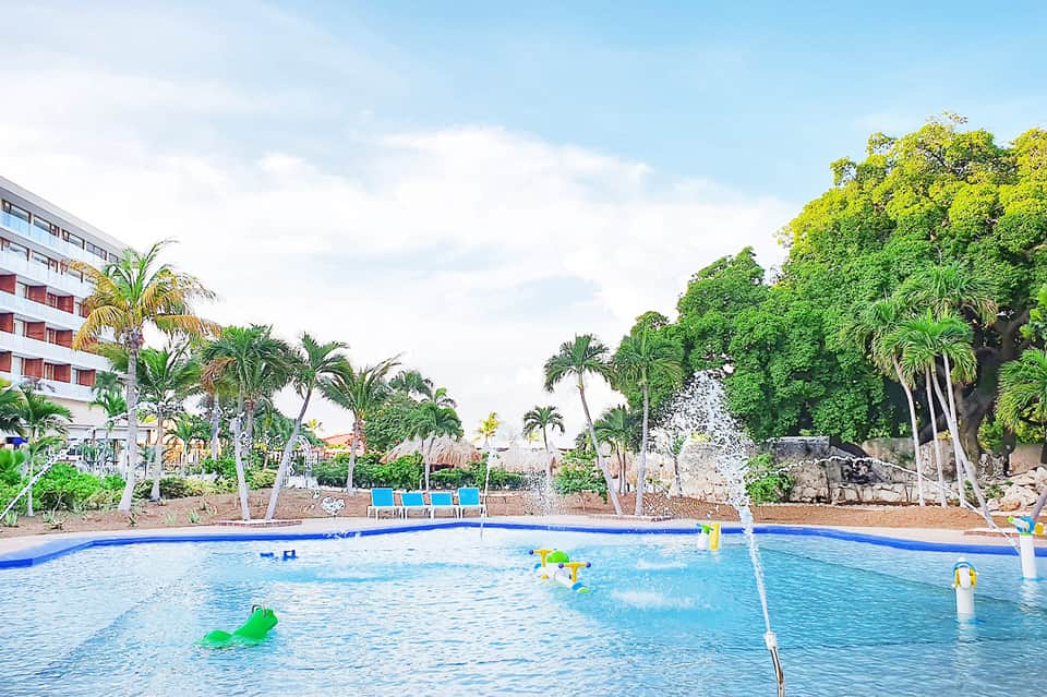 Resort pool with inflatable floats and palm trees, hotel building visible on left