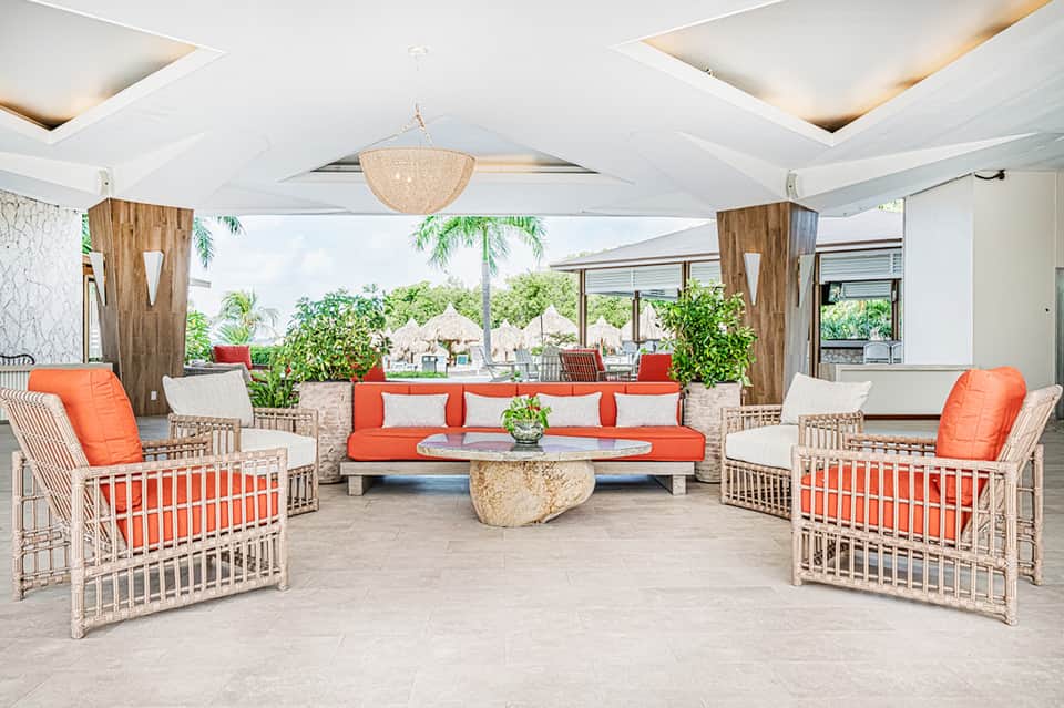 Covered resort lounge with white and orange woven chairs, coral cushions, and tropical greenery