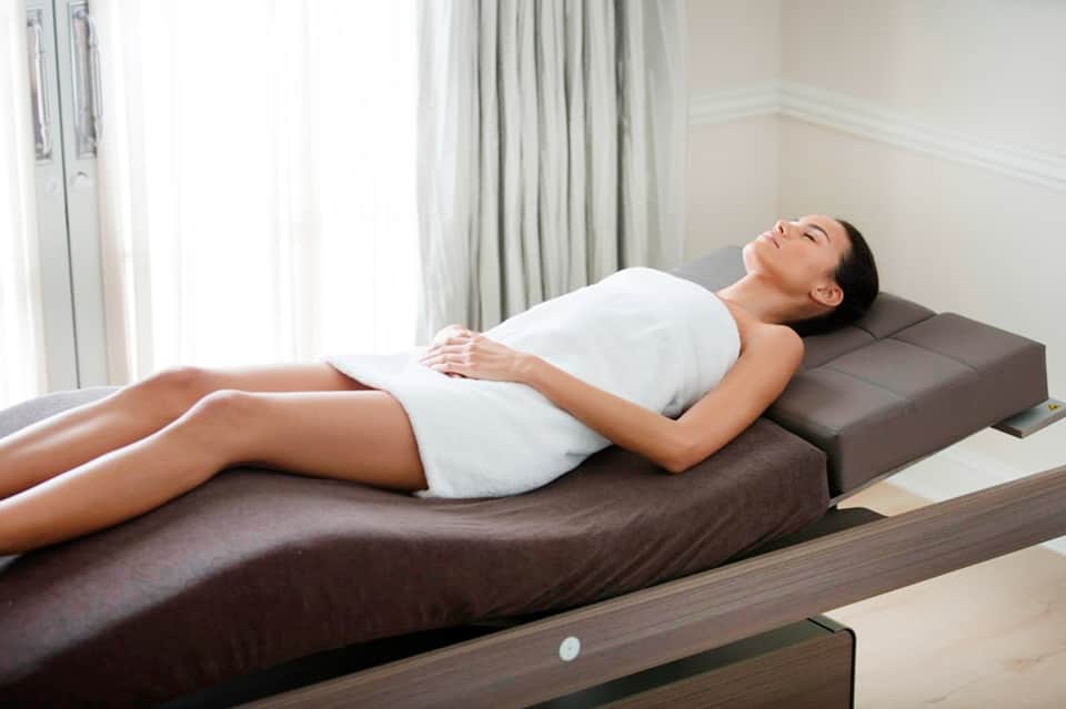 Woman in white tank top resting on a massage table with bright window lighting in background