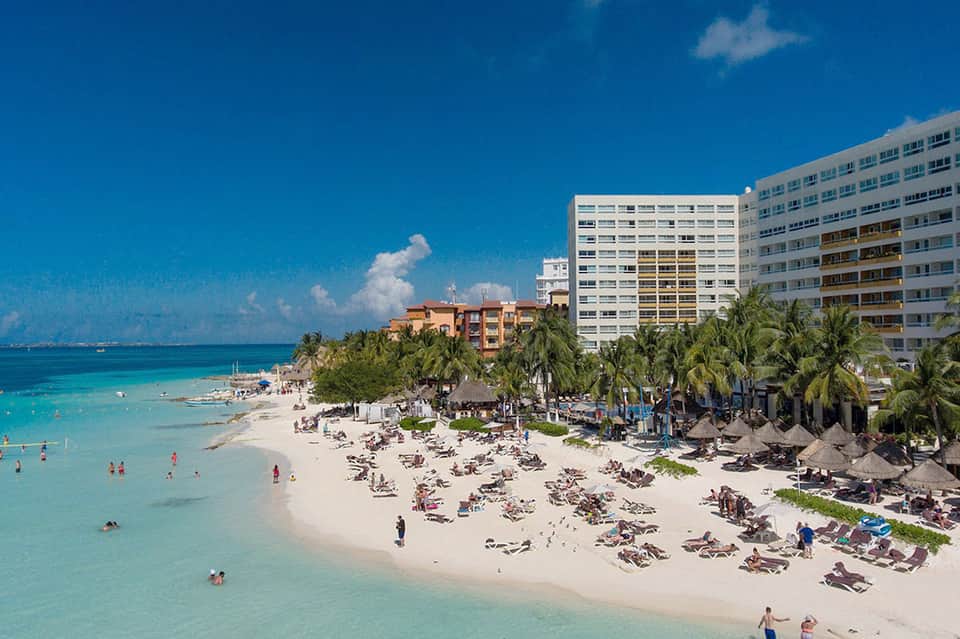 Beachfront resort with white hotel buildings, palm trees, white sand beach, and turquoise ocean