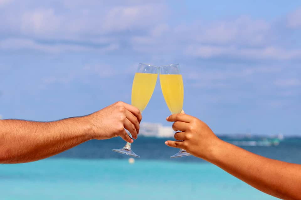 Two hands toasting champagne glasses with turquoise ocean and sailboats in background