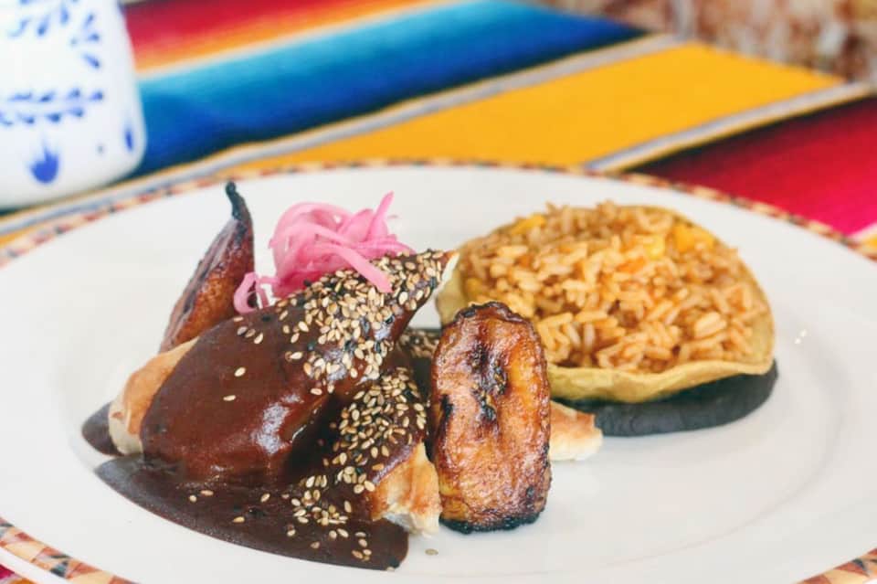Plated gourmet dish with dark sesame bun, pink pickled ginger, and side of noodles on colorful placemats