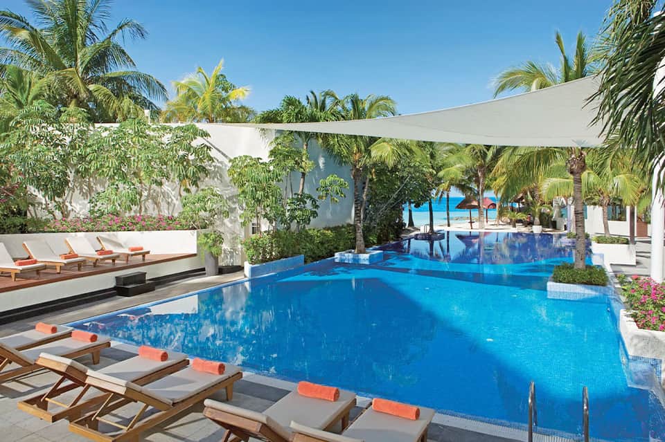 Luxury resort infinity pool with blue water, palm trees, sail shade, loungers with orange towels