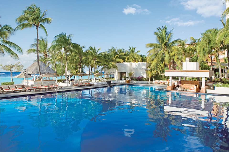 Resort infinity pool with lounge chairs, palm trees, and modern white buildings under clear sky