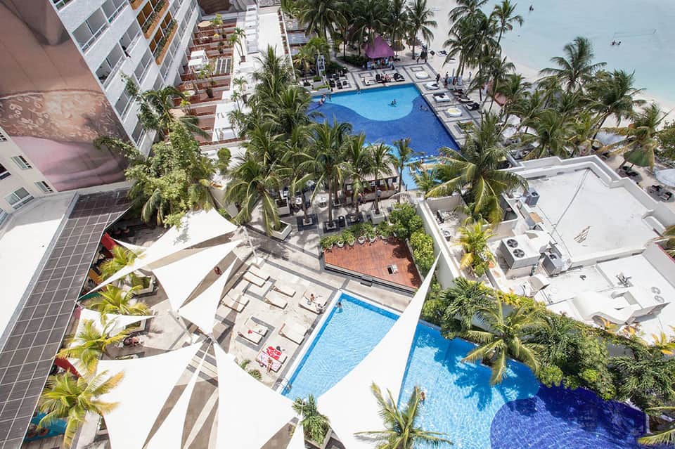 Aerial view of resort with blue pools, palm trees, beachfront, and multi-story hotel building
