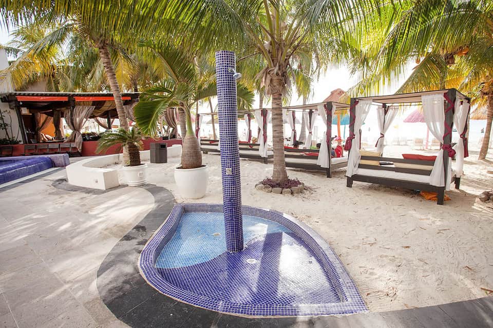 Tropical beach club with palm trees, blue mosaic fountain base, canvas cabanas, lounge seating, and sandy beach