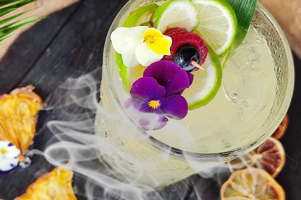 Artfully decorated cocktail with edible flowers, lime, and berries on dark surface