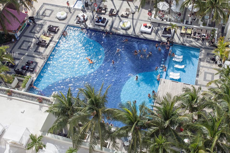 Overhead view of large resort swimming pool with sunbathers, lounge chairs, and palm trees
