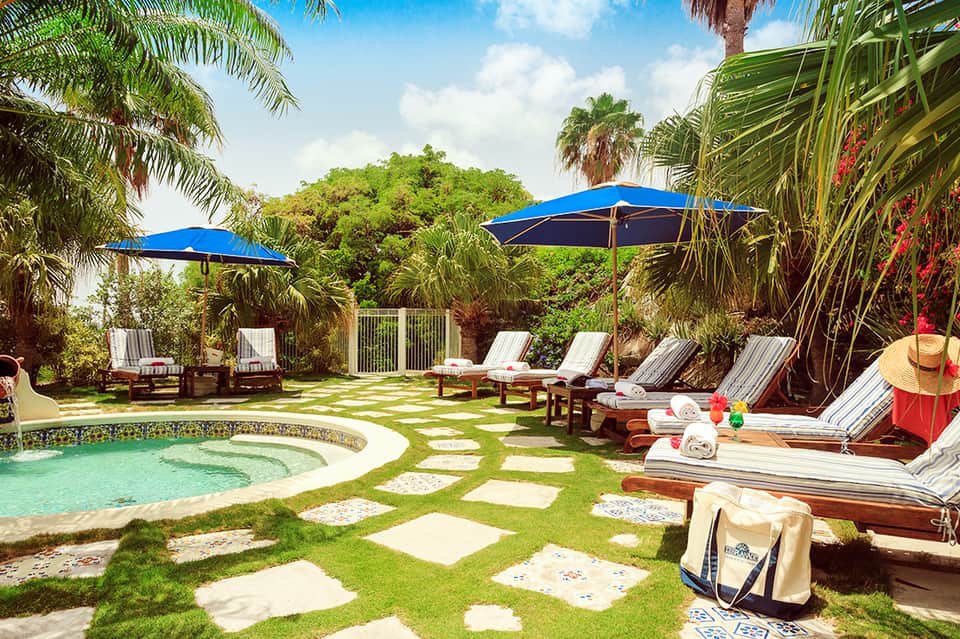 Tropical resort courtyard with spa pool, lounge chairs, blue umbrellas, palm trees, and lush landscaping