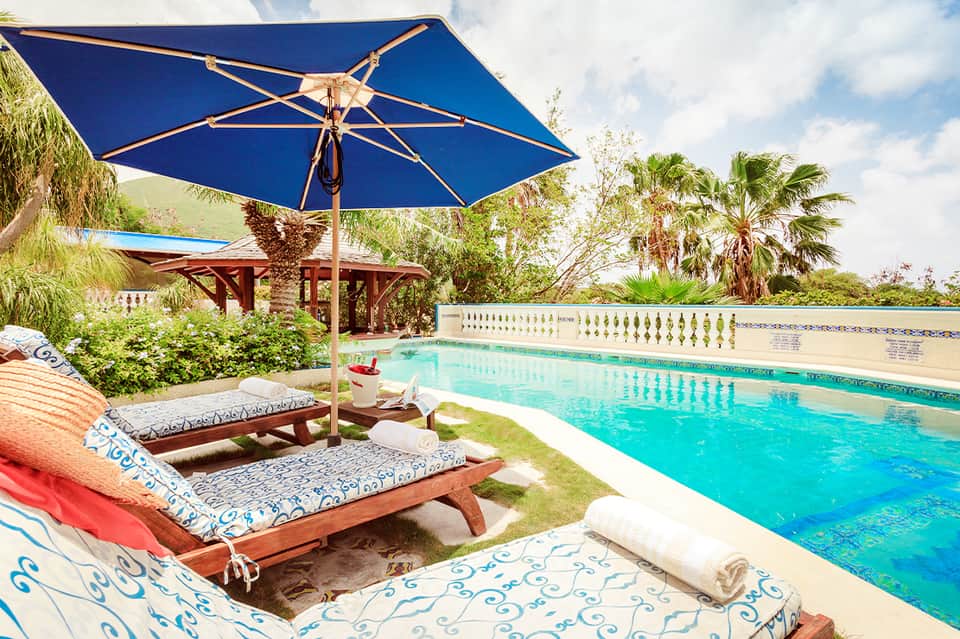 Poolside lounge chairs with blue umbrella overlooking turquoise swimming pool and landscaped gardens
