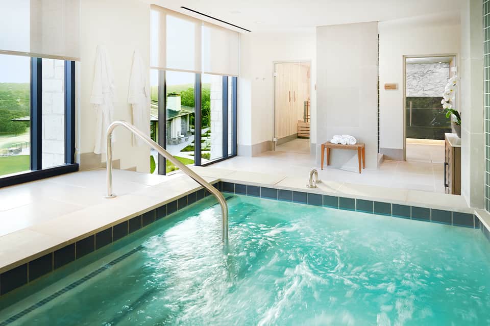 Indoor heated pool with turquoise water, white lounge area, and golf course views through glass doors