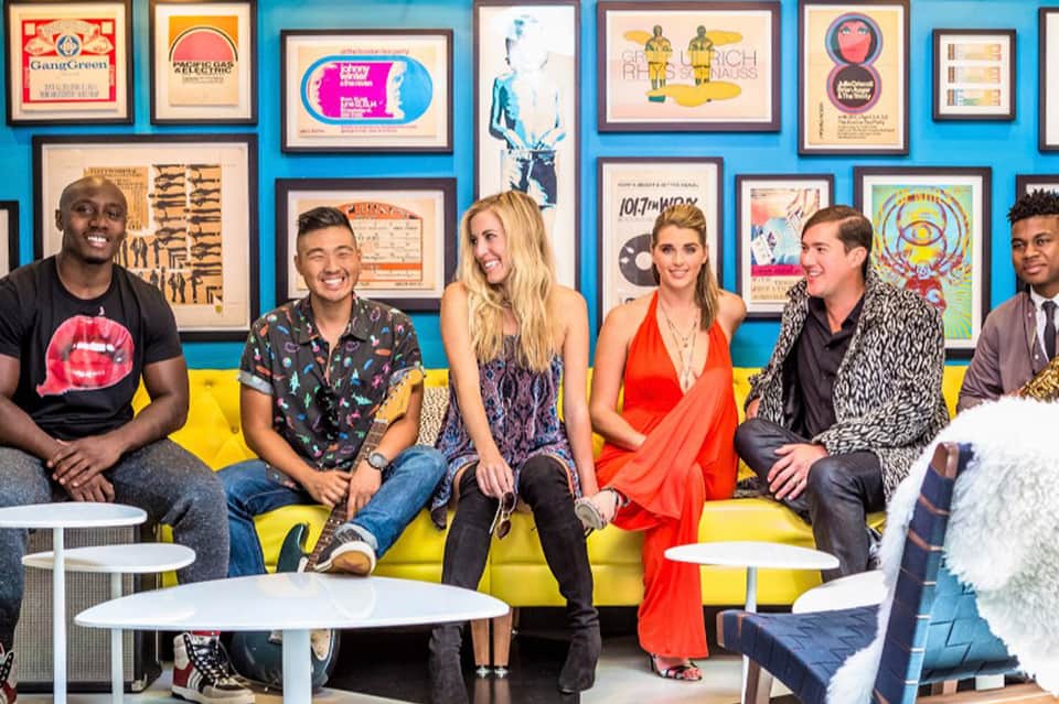 Group of six people sitting on yellow couch against blue wall with colorful framed artwork