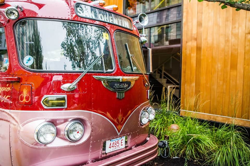 Vintage red and pink tour bus parked near wooden building and greenery