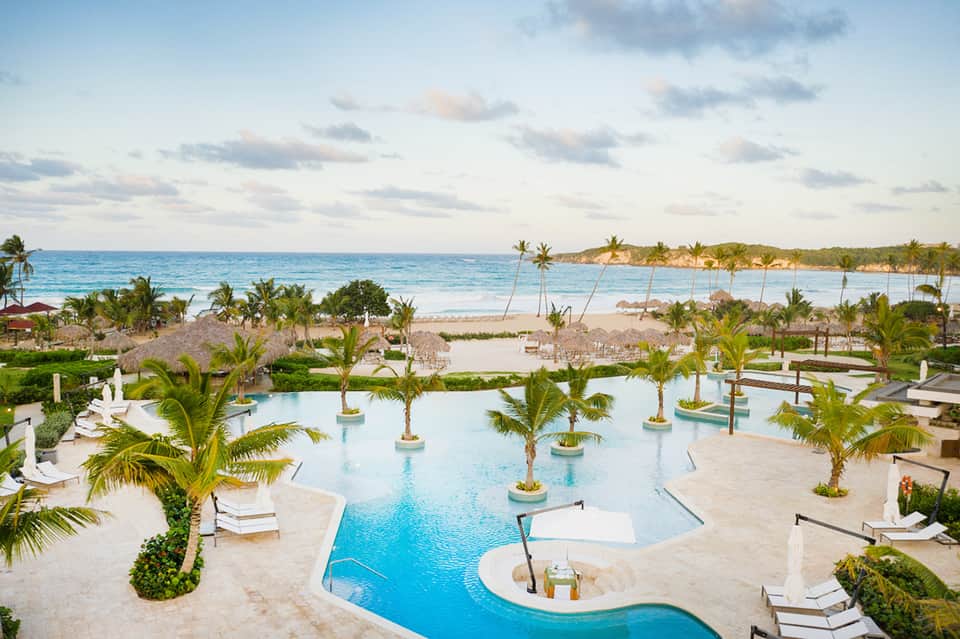 Tropical resort pool with curved design, palm trees, white loungers, and ocean beach in background