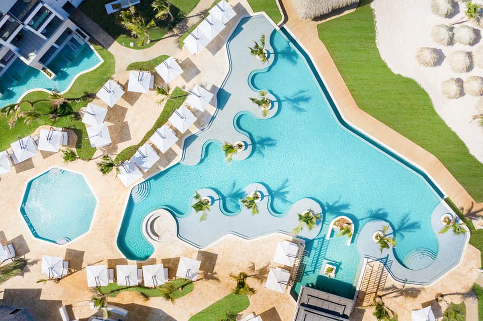 Aerial view of large resort pool complex with white loungers, green lawns, and beach access