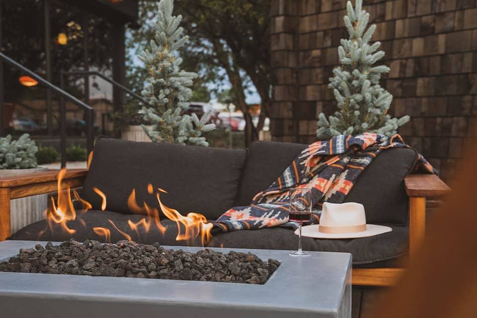 Fire pit lounge with dark sofa, blankets, and evergreen plants at dusk
