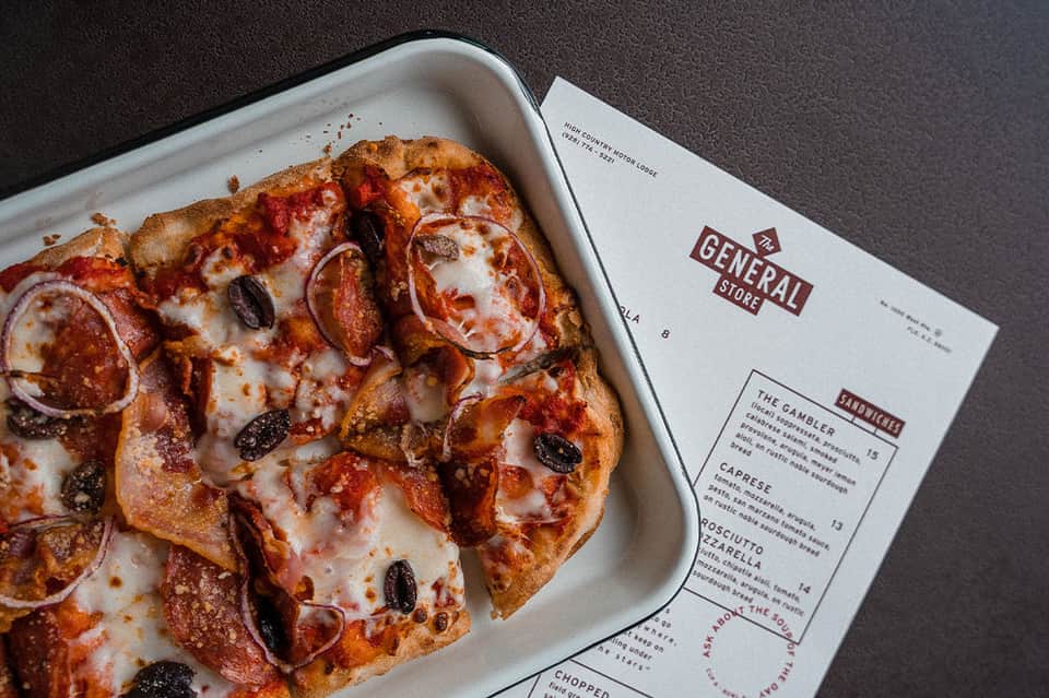 Pepperoni pizza with olives in white box next to General Store menu on dark surface