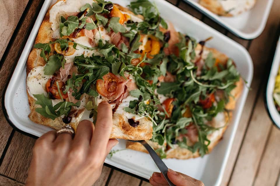 Hand reaching for gourmet pizza topped with greens, tomatoes, and prosciutto on white plate