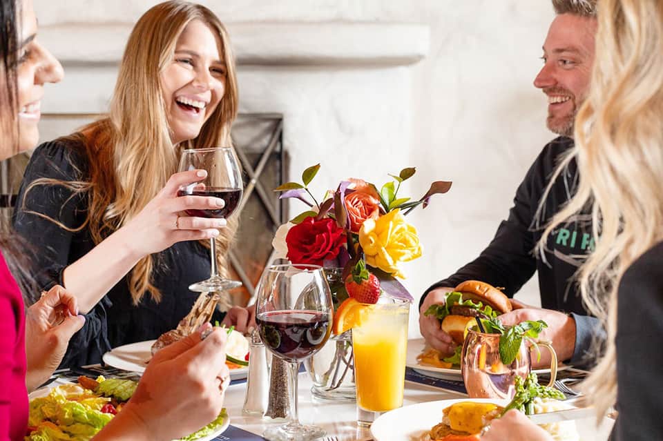 Guests dining together at elegant table with wine glasses, colorful flowers, and fresh cuisine