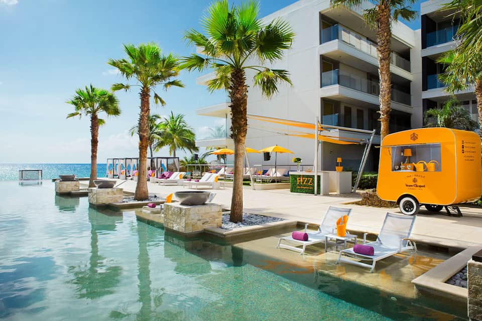 Beachfront resort with orange food cart, palm trees, and lounge chairs in shallow water