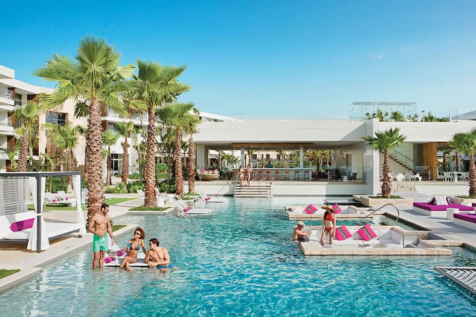 Resort pool with palm trees, modern white architecture, and guests lounging with pink accent pillows