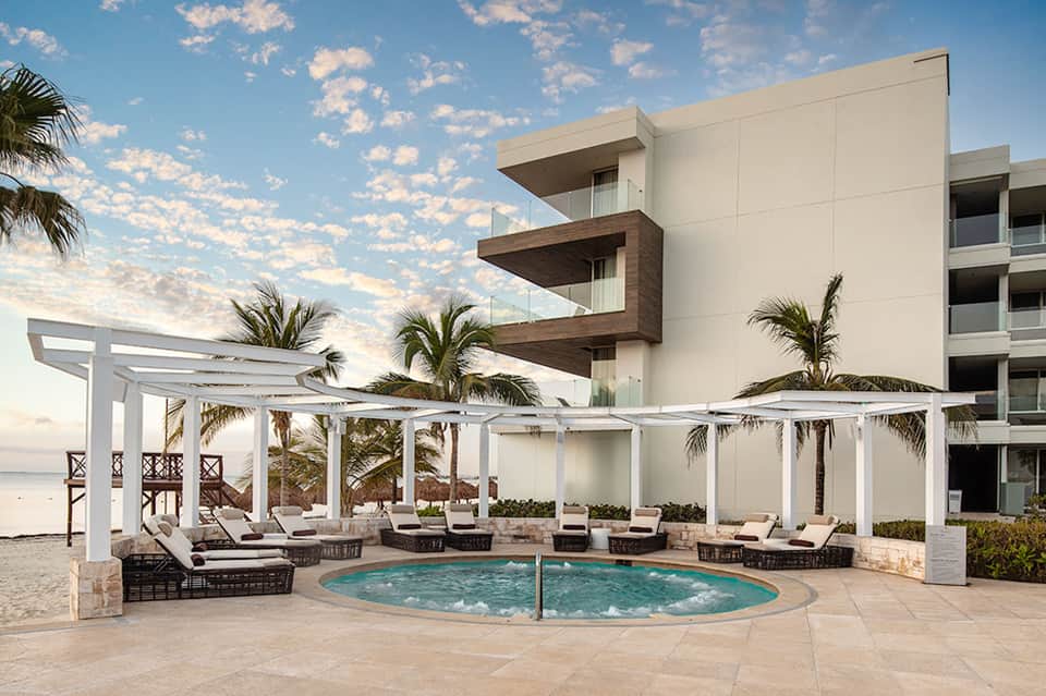 Beachfront resort with circular hot tub, white pergolas, lounge chairs, and modern building architecture