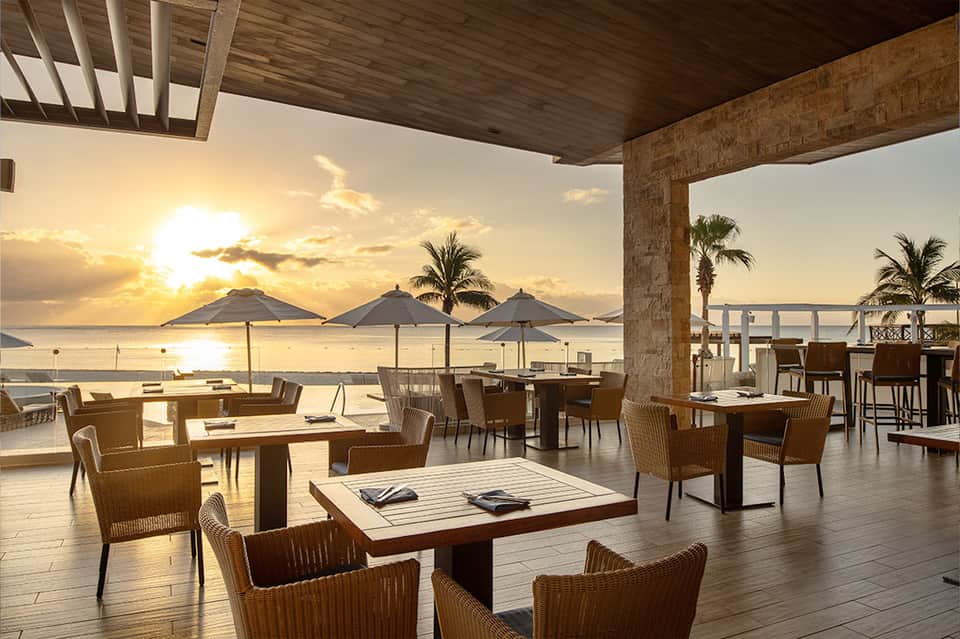 Beachfront restaurant terrace at sunset with umbrellas, wicker chairs, and ocean views