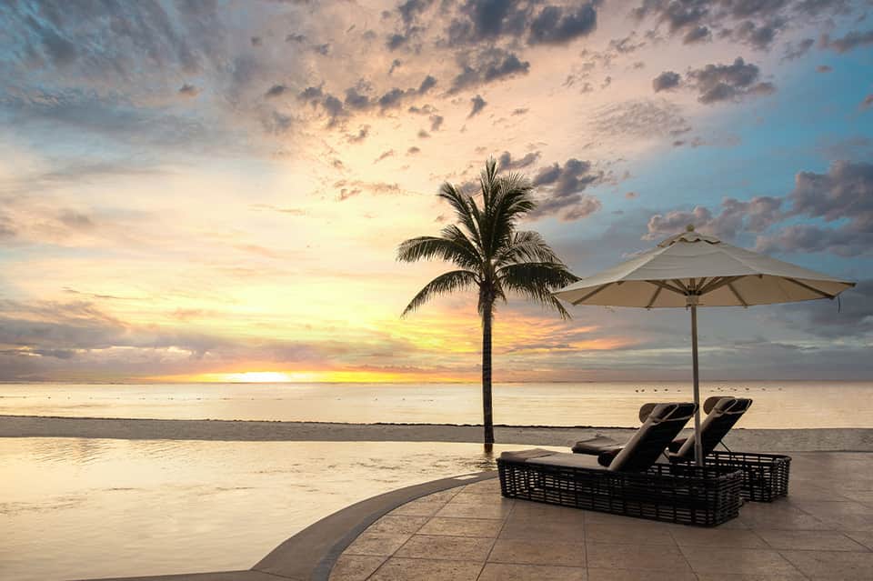 Sunset beach scene with lounge chairs under umbrella, palm tree silhouette, and golden sky reflecting on wet sand