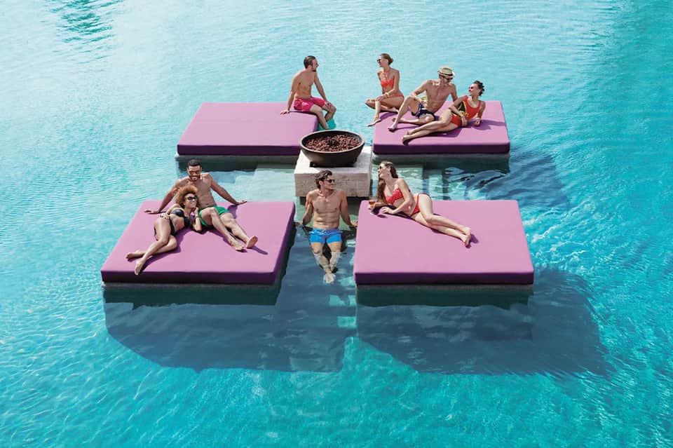 Guests floating on pink platform beds in turquoise water with central fire pit gathering area