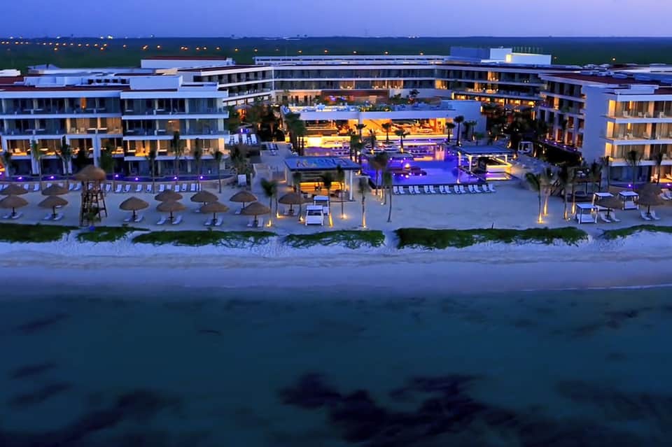 Beachfront resort at dusk with illuminated pools, loungers, umbrellas, and modern curved architecture