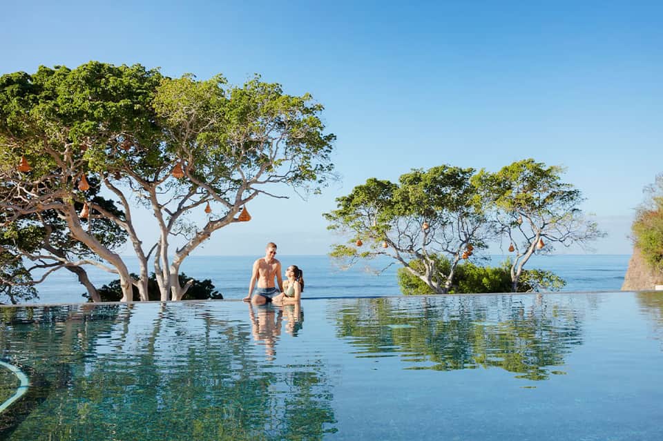 Man and woman relaxing in an infinity pool with trees and ocean horizon views