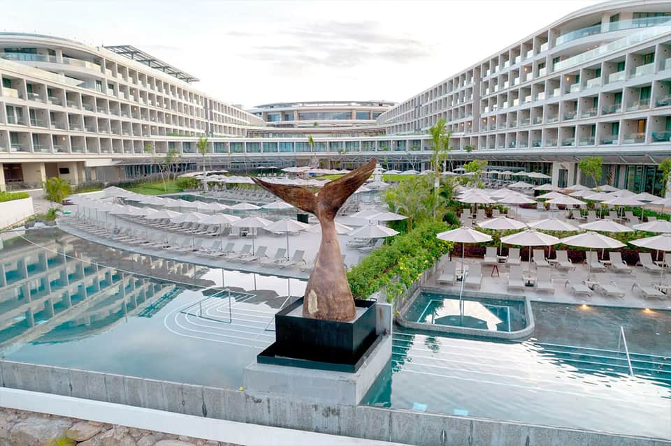 Resort courtyard with whale tail sculpture, multiple pools, loungers, and modern curved buildings