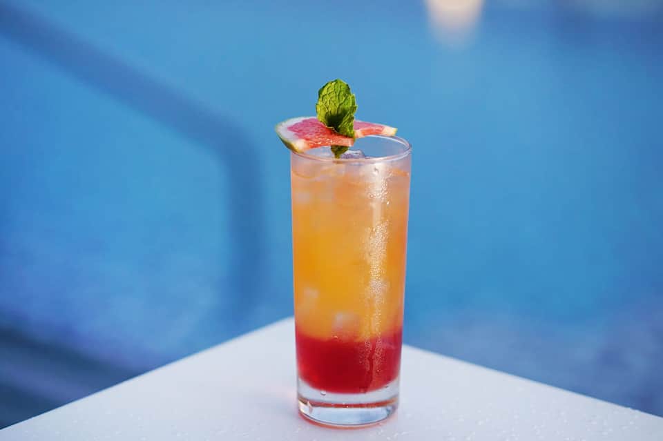 Tropical cocktail with red grenadine base, grapefruit slice and mint garnish against blue background