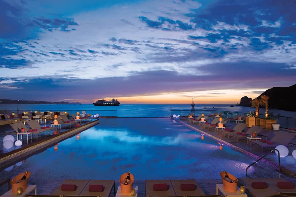 Infinity pool at twilight overlooking ocean with cruise ship, lounge seating, and lit fire bowls