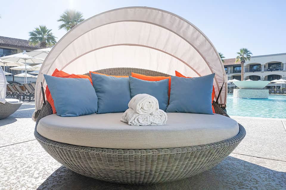 Woven daybed with blue pillows and white canopy overlooking a resort pool and palm trees