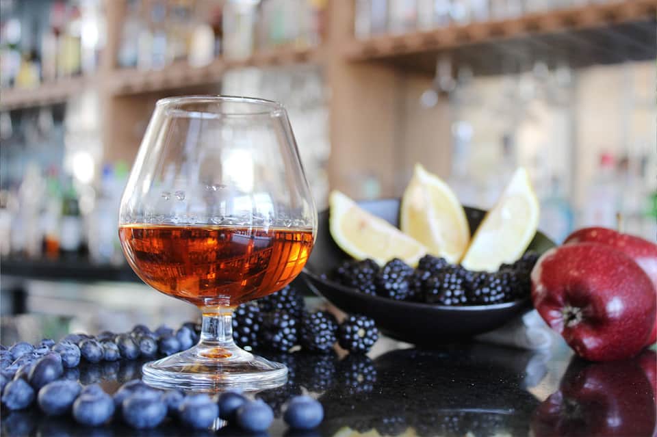 Brandy snifter with dark liquor surrounded by berries, citrus, and pomegranate at bar