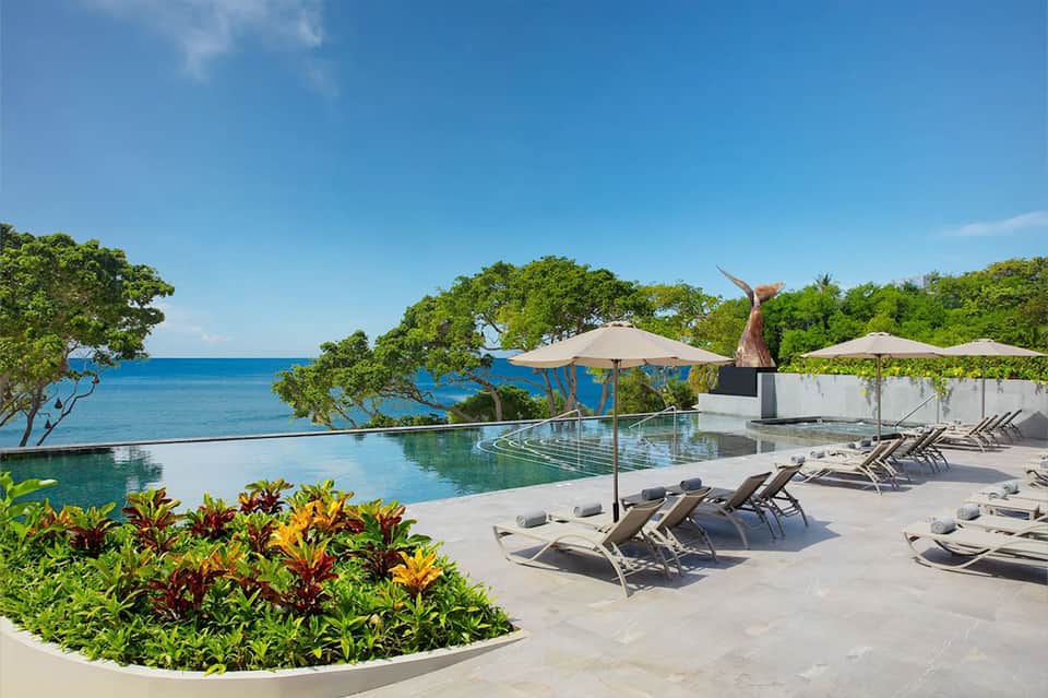 Infinity pool overlooking ocean with lounge chairs, umbrellas, and tropical landscaping