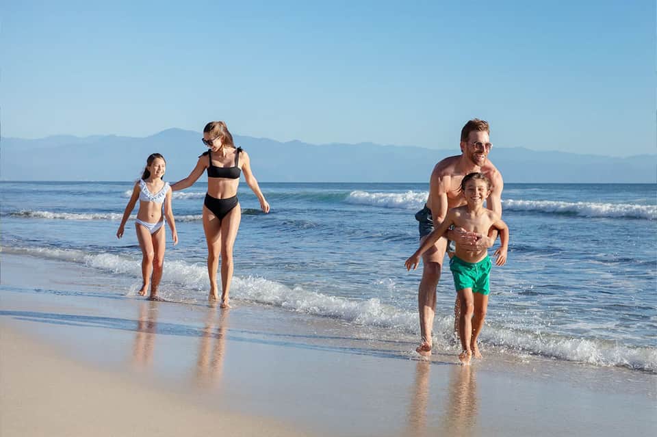 Family wading in ocean waves on sandy beach with mountains visible in background under clear blue sky