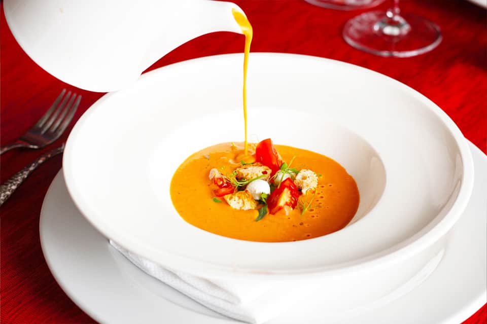 Fine dining presentation of orange soup with golden oil drizzle, red pepper garnish, and herbs on white plate