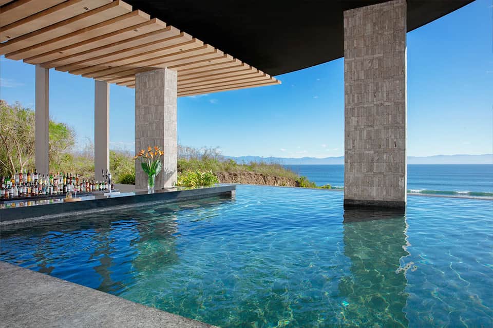 Modern infinity pool overlooking ocean, mountains, and coastline with concrete pillars and bar area