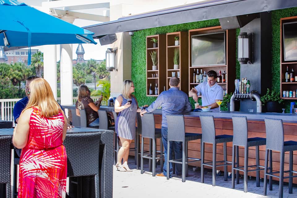 Rooftop bar with green plant wall, guests at counter with blue umbrella canopy and palm tree views