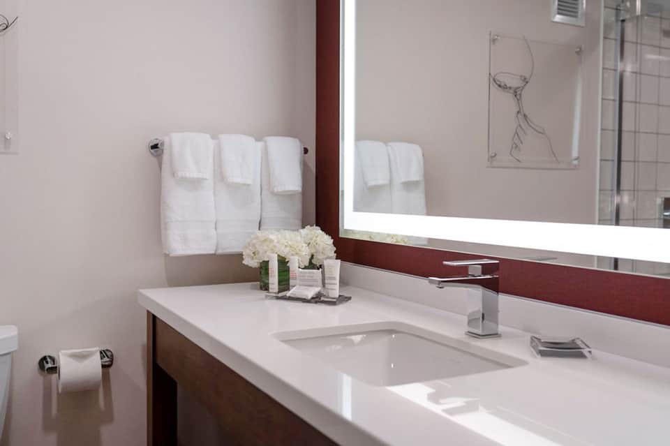 Modern hotel bathroom with white sink, illuminated mirror, dark wood vanity, and white towels