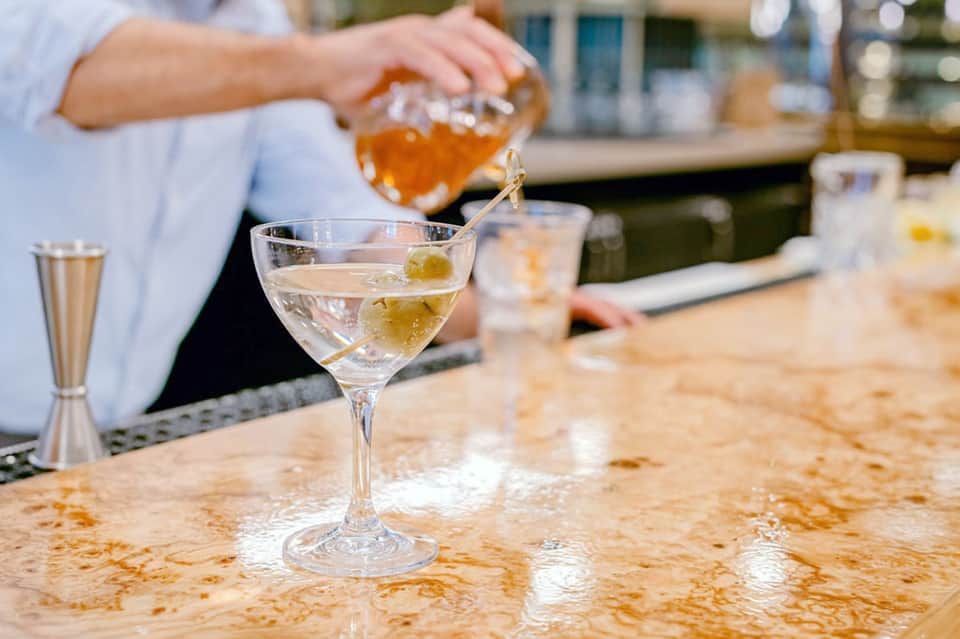 Bartender pouring amber-colored cocktail from shaker into coupe glass at outdoor bar near water